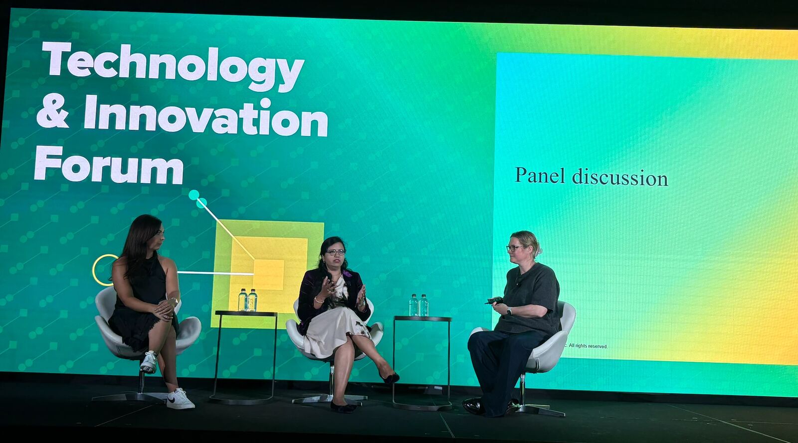 Three panelists seated in white chairs on a stage, mid conversation, in front of a large green and yellow screen that reads Technology & Innovation Forum.